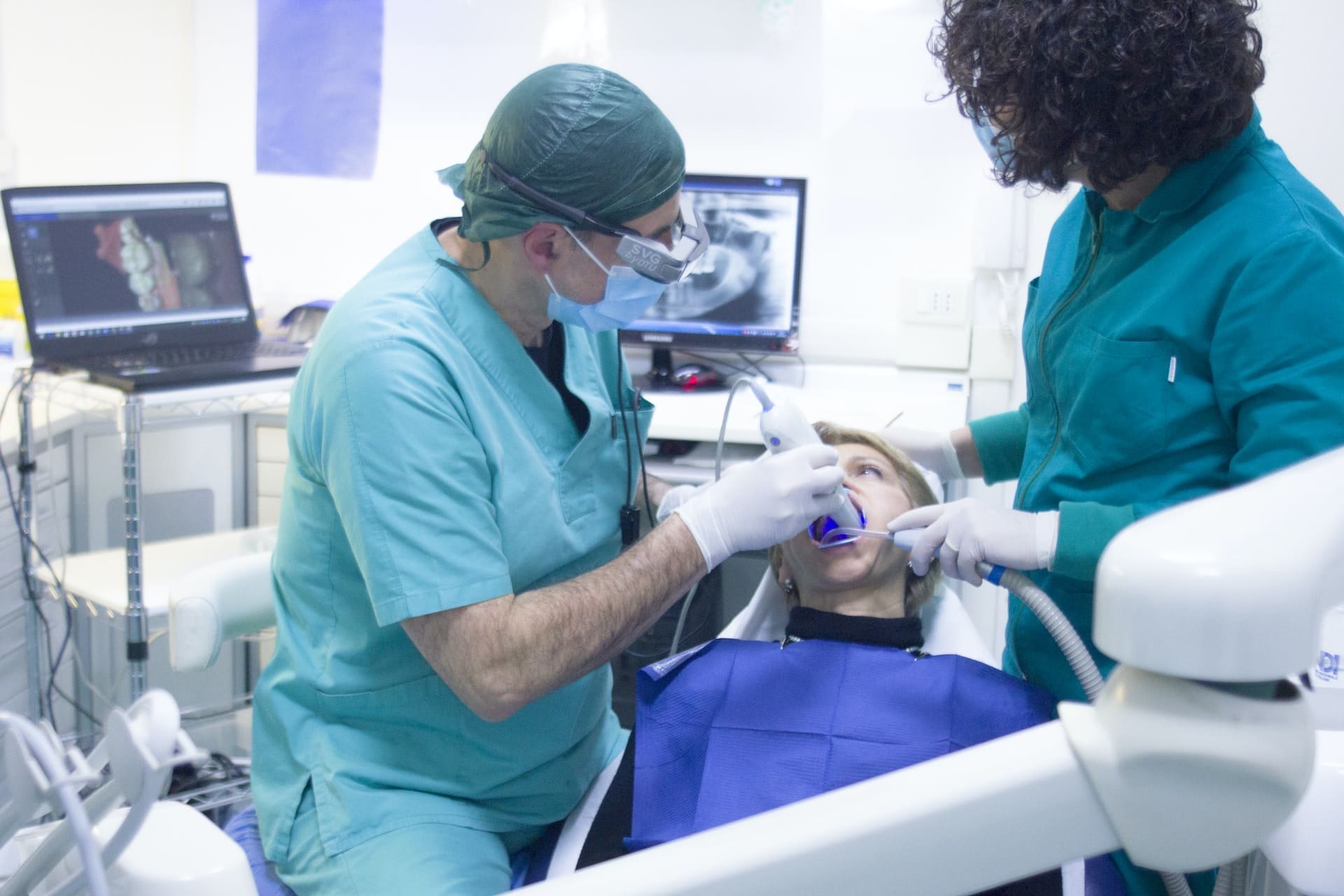 Senior dentist in surgical uniform, assisted by his assistant, placing a medical instrument into the patient's mouth during dental care.